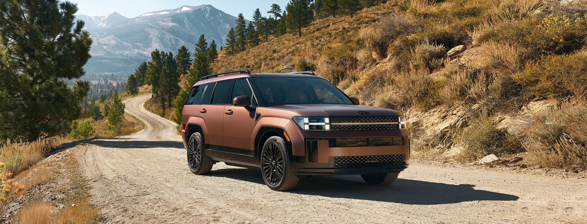2025 Hyundai Santa Fe HEV in bronze parked on a scenic dirt road surrounded by mountains and pine trees. Available now at Jerry V Honolulu Hyundai.
