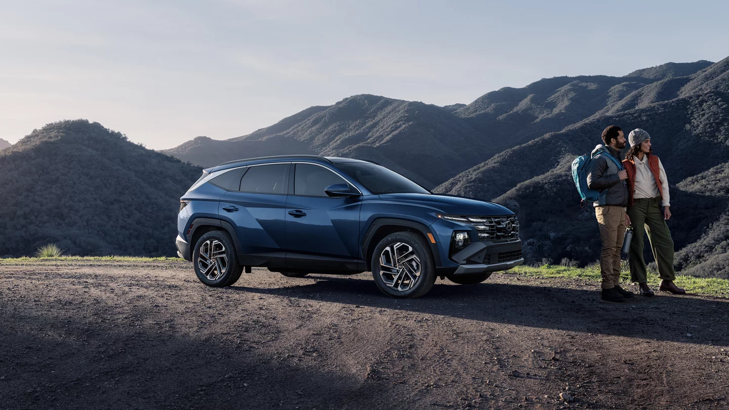 2025 Hyundai Tucson in blue parked on a dirt road with a couple standing nearby, enjoying a mountain view. Available now at Jerry V Honolulu Hyundai.
