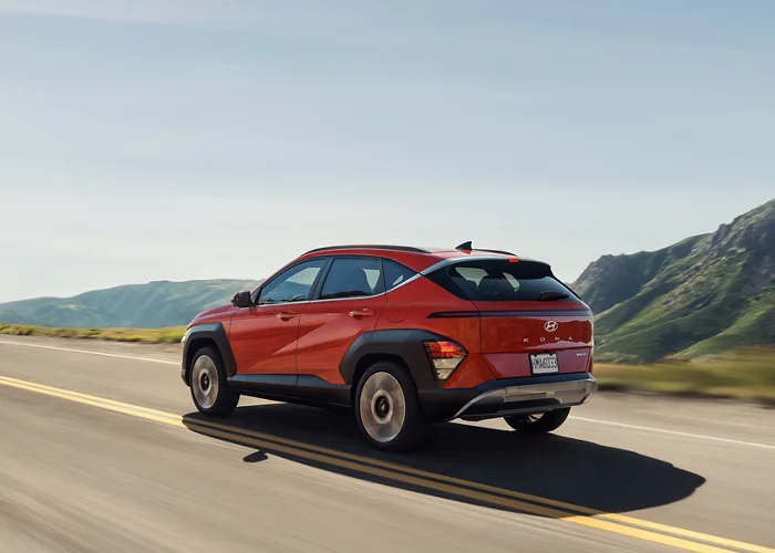 A red Hyundai Kona driving along a scenic highway surrounded by mountains, highlighting its sporty rear design.