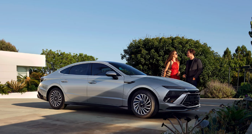Side view of a silver 2026 Hyundai Sonata parked in a modern driveway with two people standing nearby, talking next to greenery and a contemporary house.