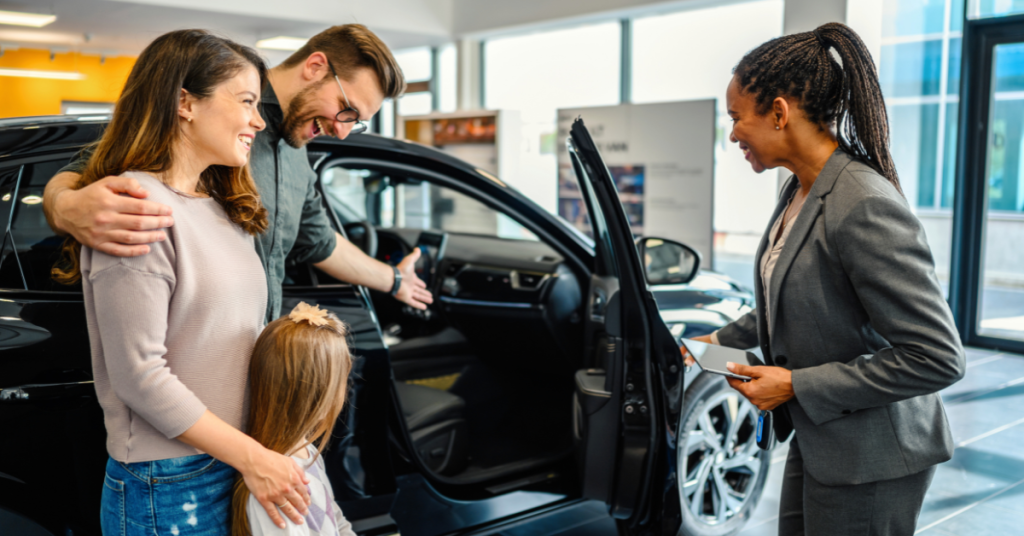 A young family with two children looks at cars in a dealership lot, discussing options with a sales representative.